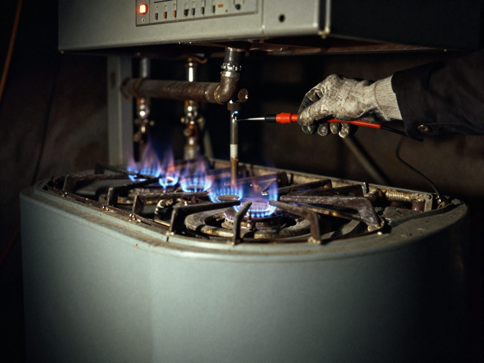 A gas heating appliance opened up in a dark basement, its row of burners lit blue, a gloved hand holding an analyzer probe down into the flame