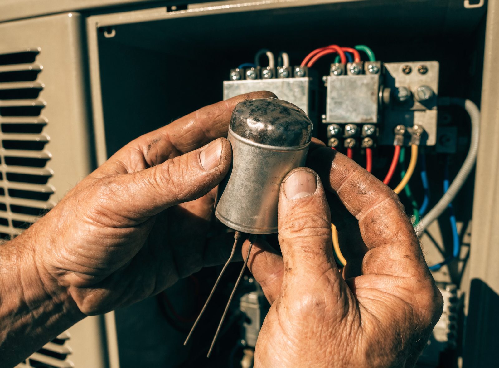 A technician's weathered hands holding a run capacitor with a swollen domed top in front of the open electrical compartment of a condenser, contactor and coloured wiring behind